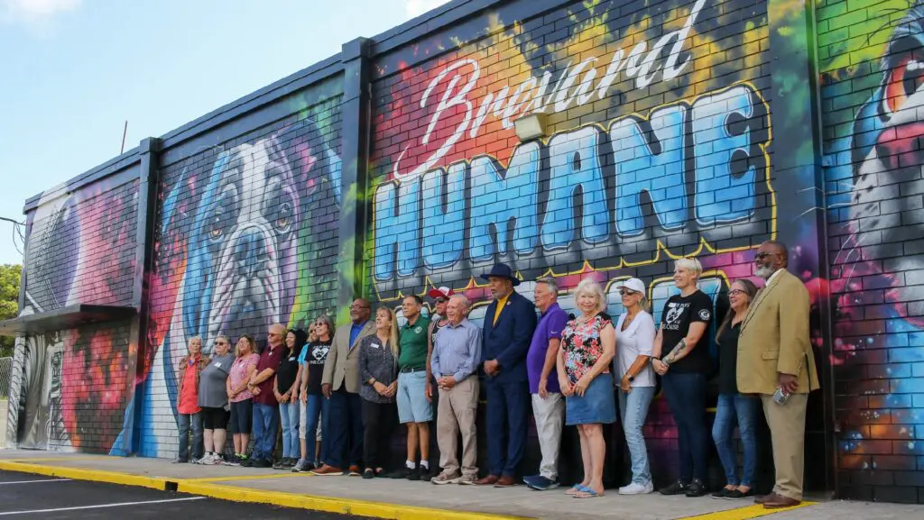 Community members gather for the mural dedication at Molly Mutt Thrift Shop in Rockledge, Florida, in front of a colorful Brevard Humane Society mural.